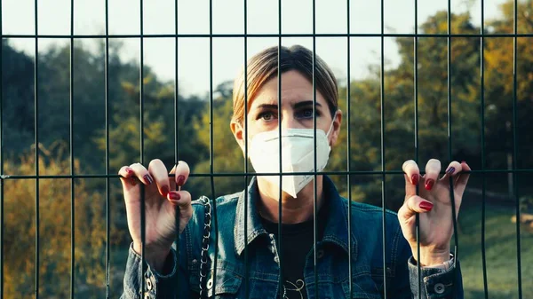 Koruyucu maskeli genç bir kadın parmaklıkların arkasından bakıyor. Karantina nedeniyle hareket edemiyor. Sınır salgından dolayı kapanmış. Yalnızlık ve yalnızlık hissi..