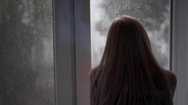 認識できない 孤独な女の子は悪天候のために家に座って 落下雨滴で窓の外を見ています 天気の良い日を待ちます バックビュー — ストック動画