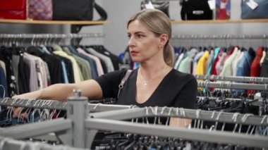 Küçük bir giyim mağazasında asılı şık kıyafetler seçen bir kadın. Sunulan giysi çeşitliliğini değerlendirir ve değerlendirir. İndirim saatlerinde alışverişe çık..