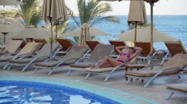 Bikinili orta yaşlı bir kadın deniz kenarındaki havuzun kenarında güneşlenir. Arka planda palmiye ağaçları olan otelde eğleniyor..