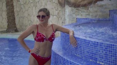 Mayo ve güneş gözlüğü takmış ince bir kadın havuzda, ferahlatıcı fıskiyelerin yanında duruyor. Otelde yaz tatili konsepti..