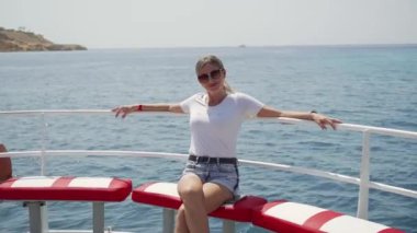 Güneş gözlüklü bir yat ya da teknenin pruvasında oturan genç bir kadın deniz yolculuğu yapıyor. Suyun üzerinde dinlenmeyi sever..
