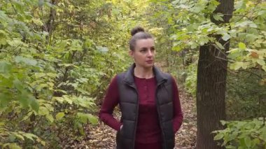 Koyu renk yelekli bir kadın sonbahar yapraklarıyla çevrili bir orman yolunda yürüyor..