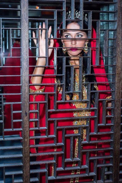 Ελκυστικό κορίτσι σε ένα κόκκινο φόρεμα ανατολίτικο στέκεται πίσω από το ξύλινο φράχτη — Φωτογραφία Αρχείου