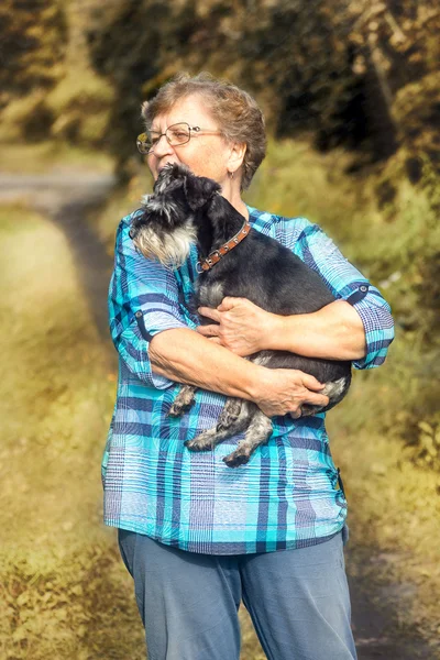 Anciana de pie sobre el fondo de la naturaleza de otoño con un perro miniatura schnauzer en las manos — Foto de Stock