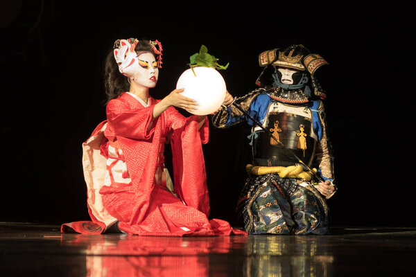 Japanese geisha in traditional kimono and fox mask holding sphere lamp and samurai warrior in armor are sitting on the knees in the dark. Traditional Japanese performance.
