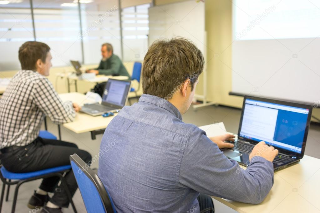People sitting at the desks and printing on the keyboards of their notebooks in a training class and the coach at the backstage
