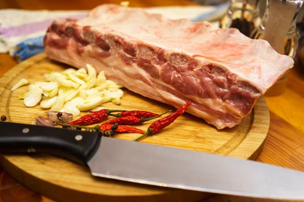 Costillas de cerdo frescas, carne marinada y preparada para asar con ajo. Colocado en una mesa de madera sobre una tabla de cortar redonda con cuchillo, pimientos fríos y ajo . —  Fotos de Stock