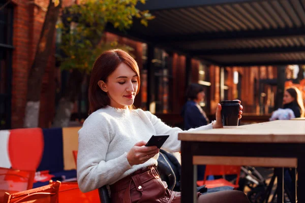 Online flört konsepti. Akıllı telefonundan mesaj okuyan ve açık kafe terasında otururken sosyal ağlarını kontrol eden gülümseyen genç bir kadın.