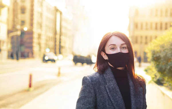 Siyah koruyucu maskeli, güneşli, boş bir sokakta, kızıl saçlı beyaz bir kadın. Coronavirus 'tan sonra canlanma. Covid salgınının sonu. Yeni normal kavram.