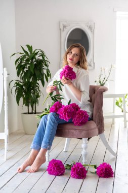 Beyaz gömlekli ve kot pantolonlu bir kadın ellerinde pembe şakayıklar, beyaz bir odada sandalyede oturuyor.