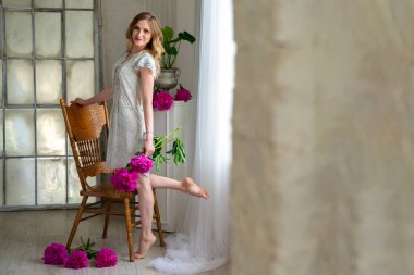 Beyaz elbiseli güzel sarışın kadın, pencerenin kenarında duruyor, elinde şakayık çiçeği tutuyor. Saç ve makyaj. Uzun bacaklar, yalınayak. Mesajın için yer aç. Boşluğu kopyala
