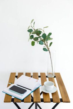 Beyaz duvarın arka planına karşı tahta masa. Okaliptüs dallı vazo, beyaz bardak ve not defterli akıllı telefon. Serbest İşyeri