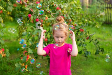 Elma bahçesindeki çocukların portresi. Pembe tişörtlü küçük kız bir elma ağacının dallarının yanında duruyor, elleriyle yüzünü kapatıyor, sabun köpüklerinden saklanıyor. Tasasız çocukluk, mutlu çocuk