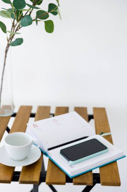 Beyaz duvarın arka planına karşı tahta masa. Okaliptüs dallı vazo, beyaz bardak ve not defterli akıllı telefon. Serbest İşyeri