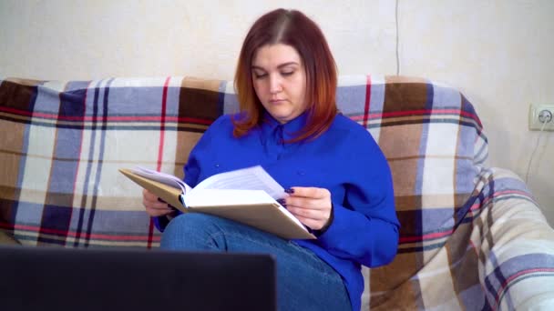 Mladá běloška sedí na gauči a čte si doma knihu — Stock video