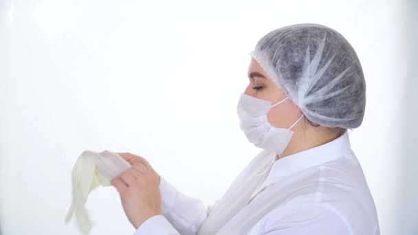 Mladá běloška, lékařka v ochranné masce a čepici, si nasazuje bílou ochrannou rukavici. Zdravotní a lékařská koncepce — Stock video