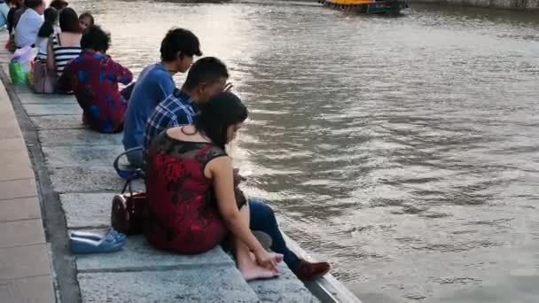 Asiáticos sentados cerca del río y el tiro de seguimiento del barco turístico en Clarke Quay en Singapur — Vídeo de stock