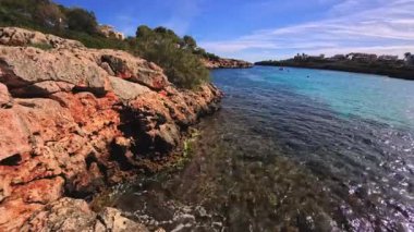 PORTOCOLOM, MALLORCA, İSPAN - 25 EKİM 2025: dalgalar kayalık kıyı şeridine hafifçe çarpar. Turkuaz su parlak güneşin altında parıldıyor. Ağır çekim için kullanılabilir..