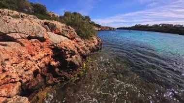 PORTOCOLOM, MALLORCA, İSPA - 25 Ekim 2025: Dalgalar kayalık kıyılara, berrak mavi sulara ve güneşli gökyüzüne çarpar. Ağır çekim için kullanılabilir..