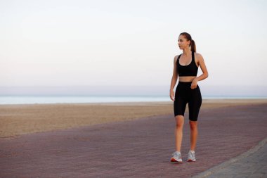 Siyah eşofmanlı ve spor ayakkabılı bir kız gün batımında deniz kenarında koşuyor.