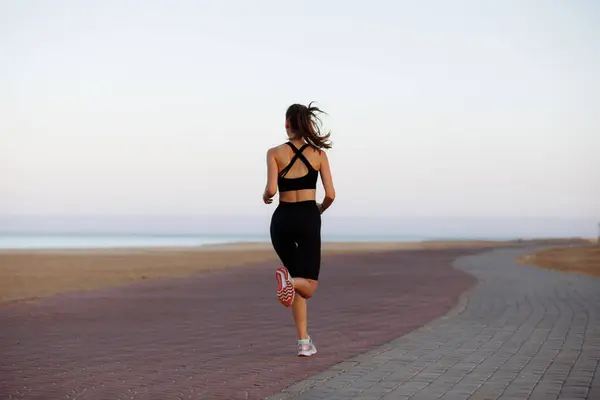 Siyah eşofmanlı ve spor ayakkabılı bir kız gün batımında deniz kenarında koşuyor.