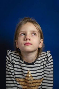 A little girl on a blue background, thinking with her eyes averted to the side, a beautiful girl with a careless hairstyle in a striped jacket with a long sleeve. think about the good and the good at a young age. clipart