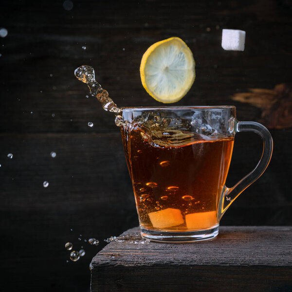 Один ломтик лимона и один кубик сахара, попадает в стакан черного крепкого чая. Замороженные брызги и капли на темном деревянном фоне.