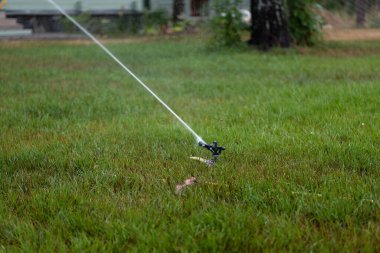 Yeşil çimenlerin üzerine monte edilmiş otomatik bir su fıskiyesinin yakından çekilmiş fotoğrafı. Fıskiyeden su püskürtülür, otomatik sulama ile bahçe bakımı yapılır..