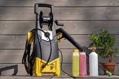 Gri bir evin arka planında araba yıkamak için mini ev tipi yüksek basınçlı yıkama makinesi ve ahşap teras. Köpük jeneratörü ve şişelerde temizlik ajansları var..