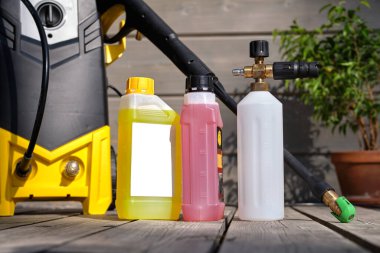 Gri bir evin arka planında araba yıkamak için mini ev tipi yüksek basınçlı yıkama makinesi ve ahşap teras. Köpük jeneratörü ve şişelerde temizlik ajansları var..