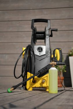 Gri bir evin arka planında araba yıkamak için mini ev tipi yüksek basınçlı yıkama makinesi ve ahşap teras. Köpük jeneratörü ve şişelerde temizlik ajansları var..