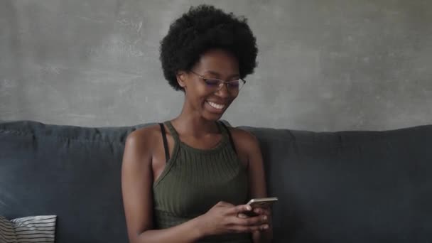 Feliz afroamericana joven mujer sentarse relajarse en acogedor sofá y feliz con el mensaje en el teléfono — Vídeos de Stock