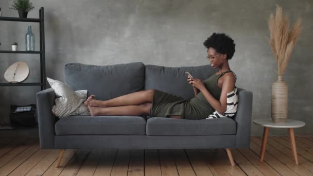 Ευτυχισμένος Αφρικανός Αμερικανός νεαρή γυναίκα κάθονται χαλαρώστε στο άνετο καναπέ ευτυχής να κινηθεί προς το νέο διαμέρισμα. Χαμογελώντας μαύρο κορίτσι στηρίζεται σε άνετο καναπέ στο σαλόνι ονειρεύεται — Αρχείο Βίντεο