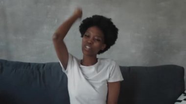 Afro-Amerikalı kız rahat bir odada kanepede telefonuyla dans ediyor.