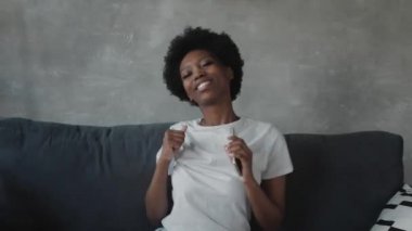 Afro-Amerikalı kız rahat bir odada kanepede telefonuyla dans ediyor.