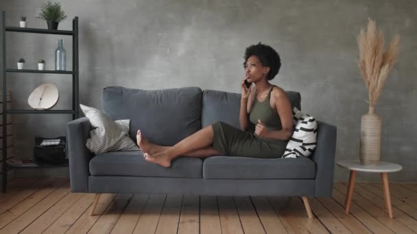 Donna afroamericana infelice che parla al telefono, ragazza arrabbiata che parla al telefono, dispositivo mobile scaricato o rotto, problema con il telefono, seduta sul divano — Video Stock