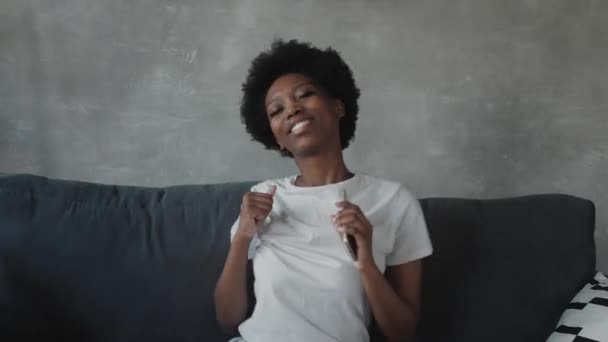 Afro-Amerikaans meisje dansen met haar telefoon op de bank in een gezellige kamer — Stockvideo