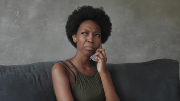 Несчастная афроамериканка разговаривает по телефону, сердитая девушка разговаривает по телефону, разряженное или сломанное мобильное устройство, проблема с телефоном, сидит на диване — стоковое видео