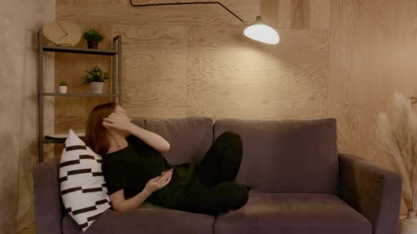 Young woman sitting on a sofa in a cozy room listening to music on headphones — Stock Video
