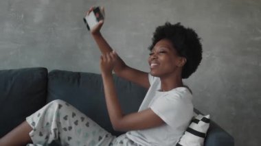 Afro-Amerikalı kız rahat bir odada kanepede telefonuyla dans ediyor.