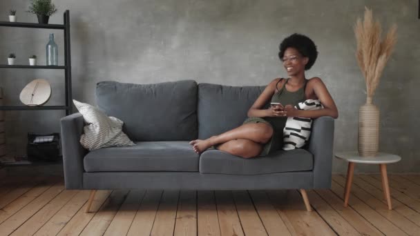 Heureuse jeune femme afro-américaine s'asseoir se détendre sur un canapé confortable et heureux avec le message au téléphone — Video