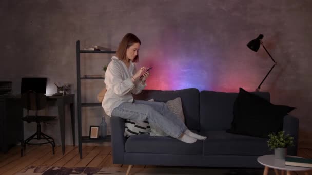 Jovem mulher asiática sentada em um sofá em um quarto acolhedor olha para o telefone — Vídeo de Stock