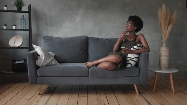 Щаслива афро-американська молода жінка сидить відпочиваючи на затишному дивані і щаслива з повідомленням по телефону. — стокове відео