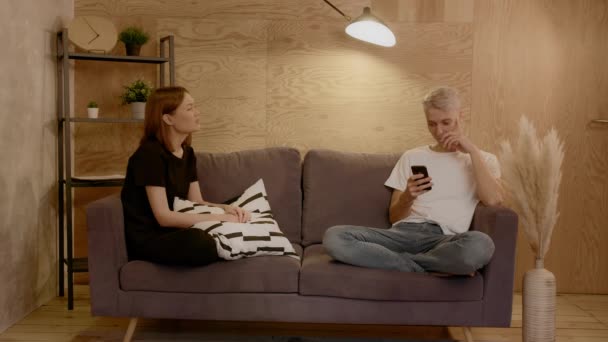 Et ungt par sidder på sofaen. Pigen prøver at tale med fyren. Fyren reagerer ikke på pigen. – Stock-video