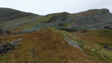 Ffestiniog, Galler 'deki Llyn Cwmorthin üzerinde dinamik FPV insansız hava aracı videosu. Sahne, engebeli, dağlık kayalık manzarayı dramatik, karamsar ve bulutlu bir gökyüzü altında yakalar..