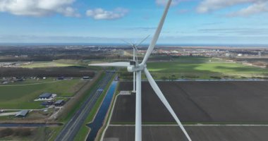 Arka planda kırsal bir manzara, otoyol ve kanal ile temiz ve yenilenebilir elektrik üretmek için dönen bıçakları büyük bir rüzgar türbininin yanında uçan dramatik hava perspektifi