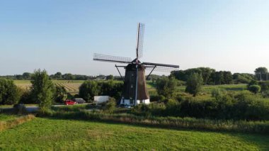Uçan pervaneli tarihi bir Alman yel değirmeninin havadan görünüşü, açık mavi gökyüzünün altında yemyeşil tarlalar ve tarım alanlarının ortasında duruyor, kırsal Hollanda mirasının ve tarımın özünü yakalıyor.