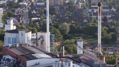 Evleri ve yeşil ağaçları olan ve kentsel gelişimi ön plana çıkaran geniş bir yerleşim alanının yanındaki bacaları ve bacaları olan büyük bir endüstriyel kompleksin havadan görünüşü