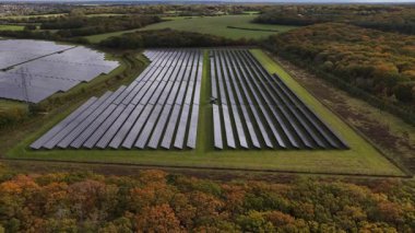 Geniş güneş çiftliği, güneş ışığından temiz ve sürdürülebilir elektrik üreten sıra sıra fotovoltaik paneller, verimli yeşil kırsal ve sonbahar ormanları ile çevrili bulutlu bir gökyüzü altında